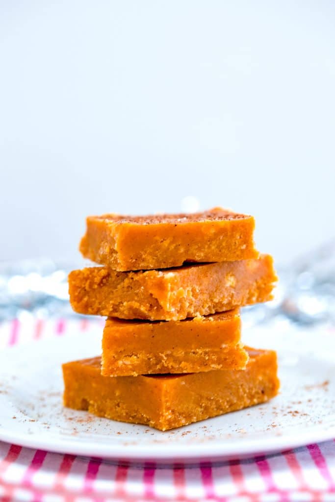 Head-on view of pumpkin fudge squares stacked on a white plate on a pink and red towel with pumpkin spice sprinkled around