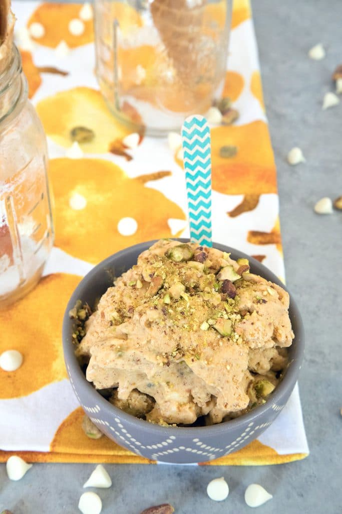 Overhead view of a bowl of pumpkin ice cream with pistachio swirl with a wooden spoon in it and surrounded by white chocolate chips