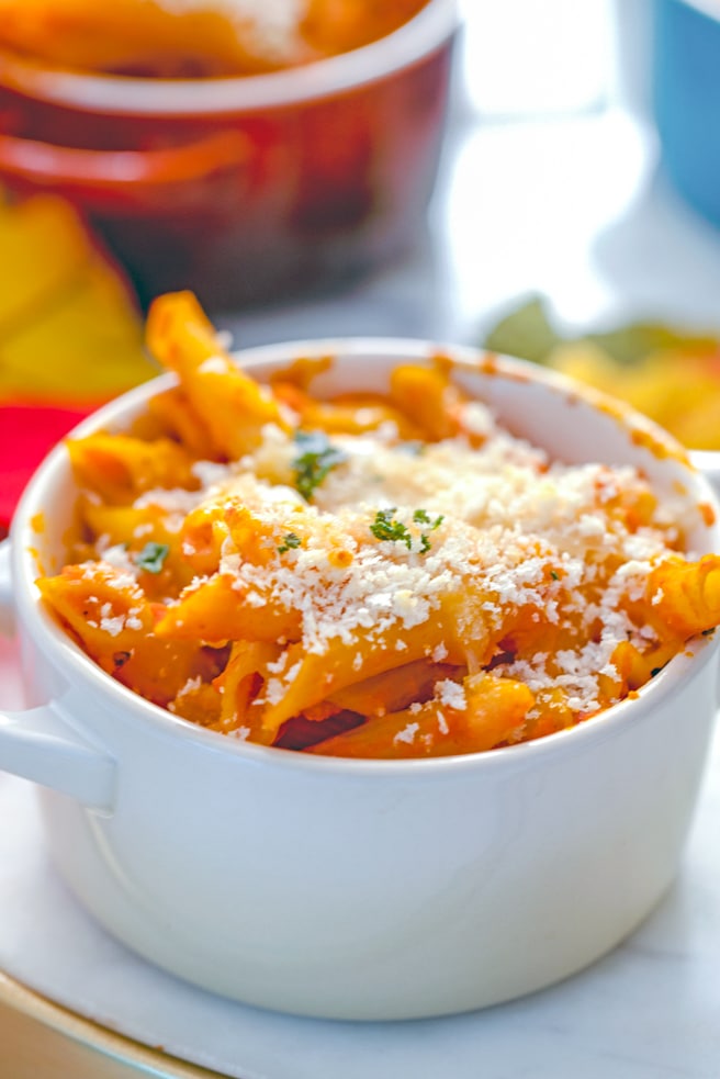 Close-up head-on view of white ramekin filled with pumpkin mac and cheese with more ramekins and fall leaves in the background