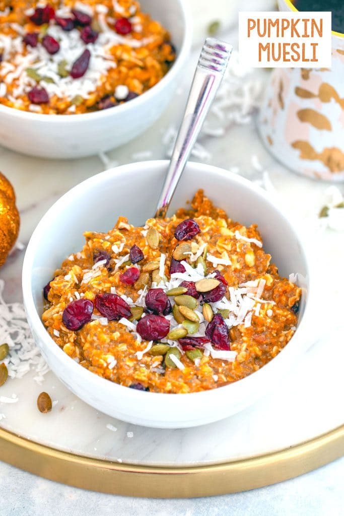 Overhead view of a white bowl of pumpkin muesli topped with dried cranberries, coconut, and pumpkin seeds on a marbled trivet with second bowl in the background, along with a cup of coffee, coconut sprinkled around, and recipe title at the top of the image