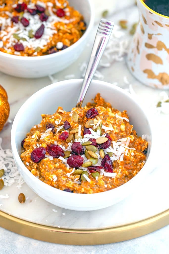 Overhead view of a white bowl of pumpkin muesli topped with dried cranberries, coconut, and pumpkin seeds on a marbled trivet with second bowl in the background, along with a cup of coffee, and coconut sprinkled around