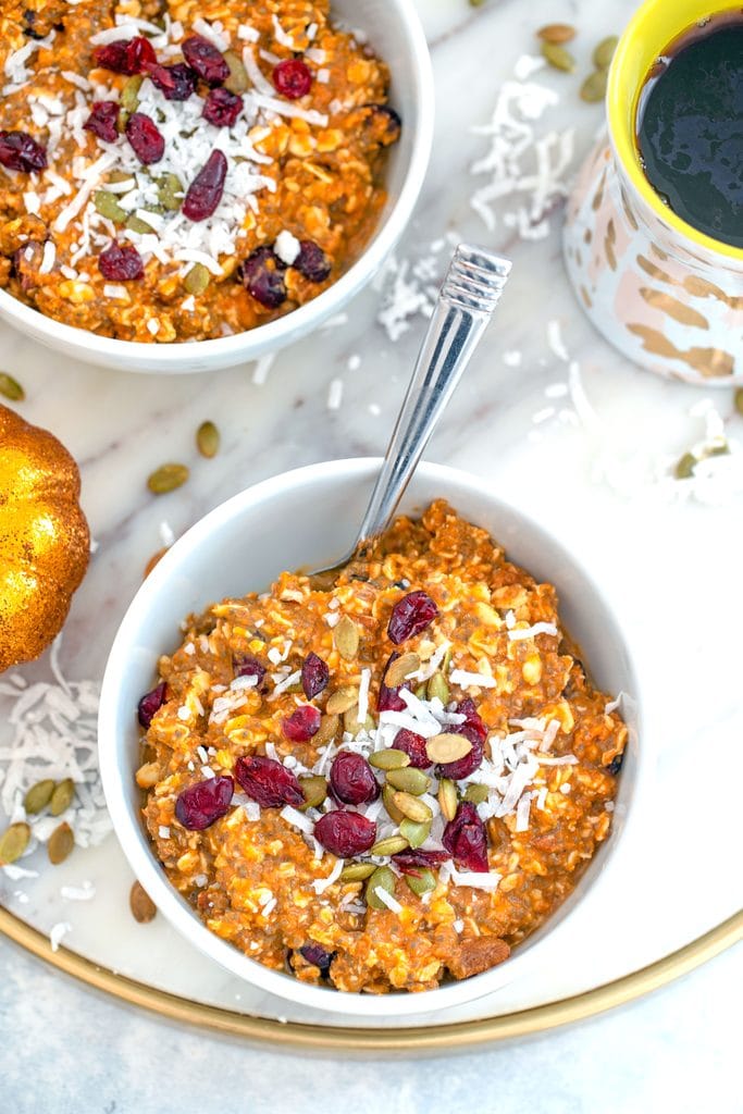 Bird's eye view of two bowls of pumpkin muesli on a marble trivet with coconut and pumpkin seeds sprinkled around and a cup of coffee in the background