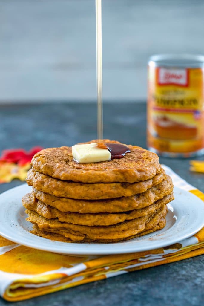 Head-on view of a stack of pumpkin pancakes on a white plate with a pat of butter and maple syrup being poured over the top