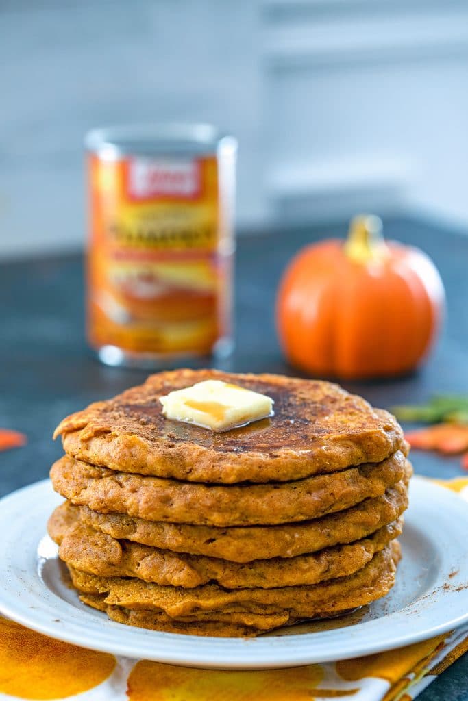 Head-on view of a stack of pumpkin pancakes with a pat of butter on top and a can of pumpkin puree and pumpkin in the background