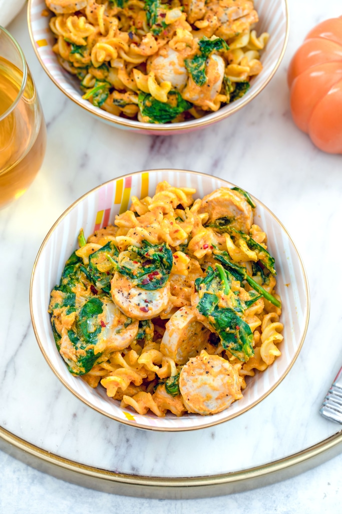 Bird's eye view of two bowls of pumpkin pasta with chicken sausage and spinach on a marble tray with glass of white wine and pumpkin in the background