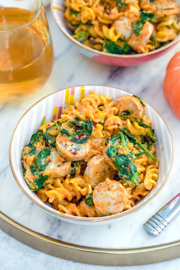 Overhead view of a bowl of pumpkin pasta with chicken sausage with spinach on a marble tray with second bowl of pasta and glass of white wine in background