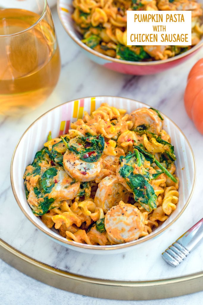 Overhead view of a bowl of pumpkin pasta with chicken sausage with spinach on a marble tray with second bowl of pasta and glass of white wine in background with recipe title at top