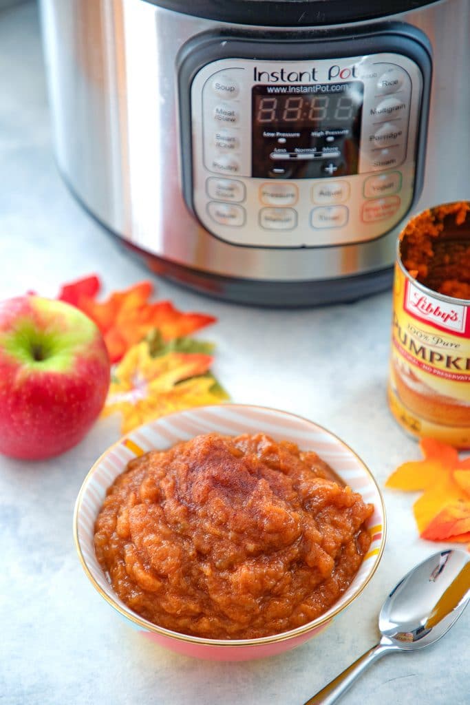 Head-on view of a bowl of pumpkin pie applesauce with a spoon, apple, can of pumpkin puree, leaves, and Instant Pot in the background