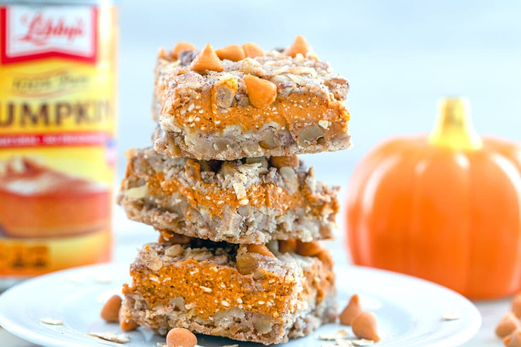 Landscape photo with closeup of three pumpkin pie bars stacked on each other on a white plate with a can of pumpkin puree and a small ceramic pumpkin in the background
