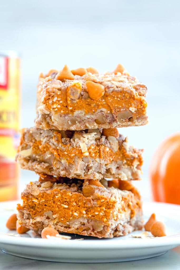 Head-on closeup view of stack of three pumpkin pie bars on a white plate with butterscotch chips and oats on plate and can of pumpkin puree in background