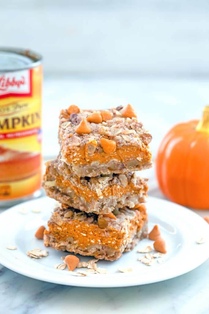 Head-on view from a distance of three pumpkin pie bars stacked on a white plate and surrounded with butterscotch chips and oats with a can of pumpkin puree and small pumpkin in the background