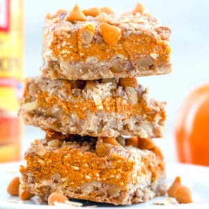 Head-on closeup view of stack of three pumpkin pie bars on a white plate with butterscotch chips and oats on plate and can of pumpkin puree in background