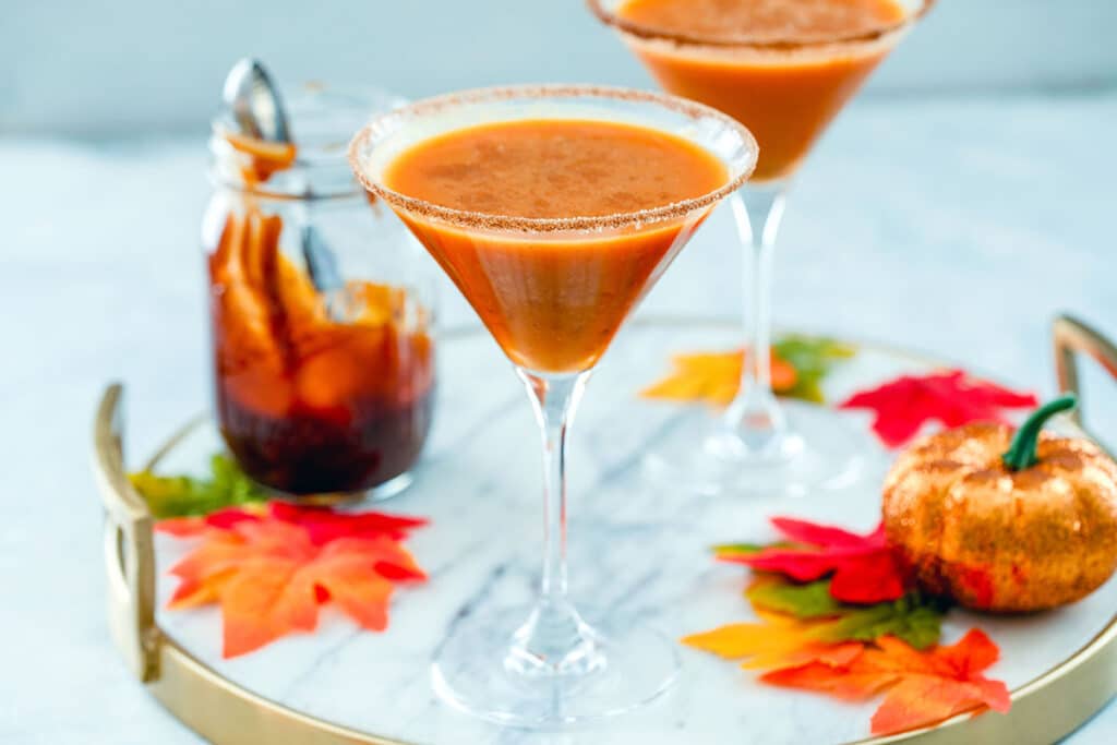 Landscape photo of two pumpkin pie martinis on a marble surface with jar of caramel sauce in the background, along with maple leaves and a small glitter pumpkin
