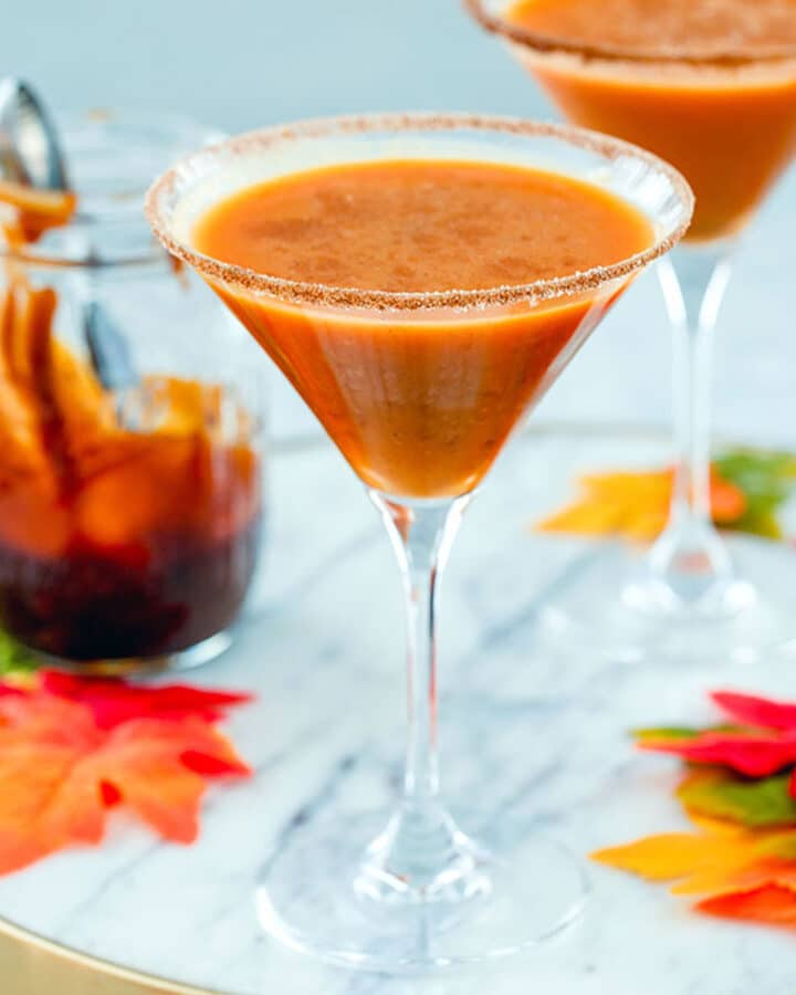 Head-on close-up view of a pumpkin pie martini on a marble surface with a second one, maple leaves, and a jar of caramel sauce in the background
