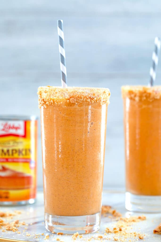 Head-on close-up view of a orange pumpkin pie milkshake with graham cracker rim and grrey and white straw with second milkshake and can of pumpkin in the background