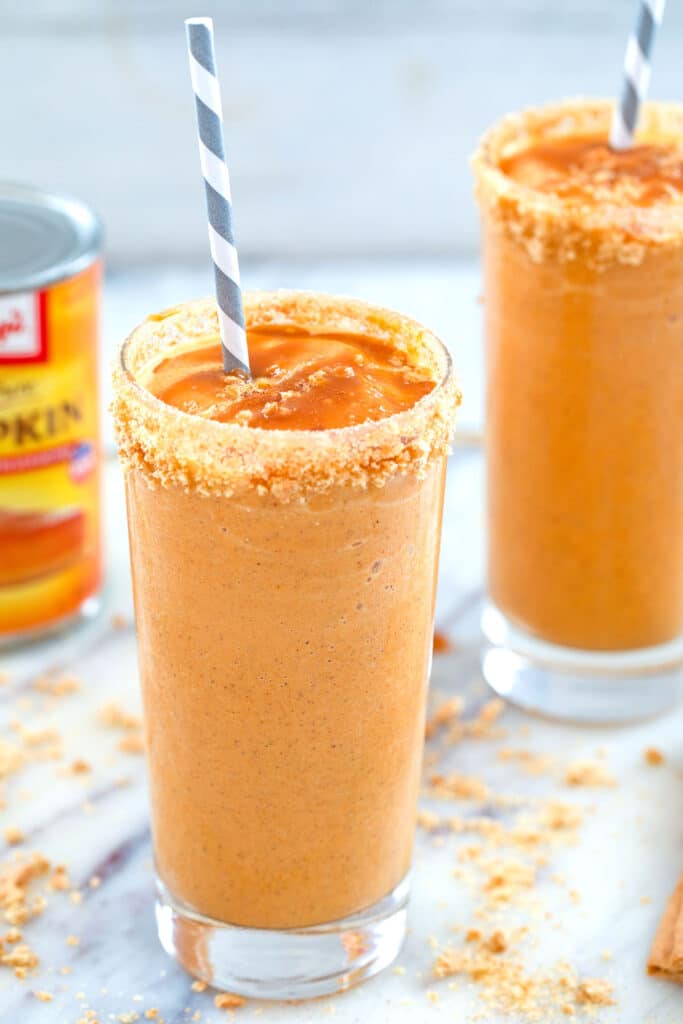 View of pumpkin pie milkshake in a graham cracker rimmed glass with caramel drizzle and grey and white straw with can of pumpkin and second milkshake in the background