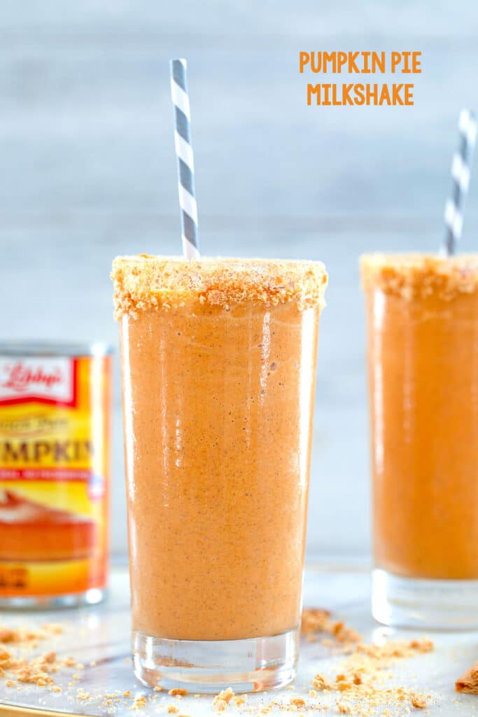 Head-on close-up view of a orange pumpkin pie milkshake with graham cracker rim and grrey and white straw with second milkshake and can of pumpkin in the background with recipe title at top