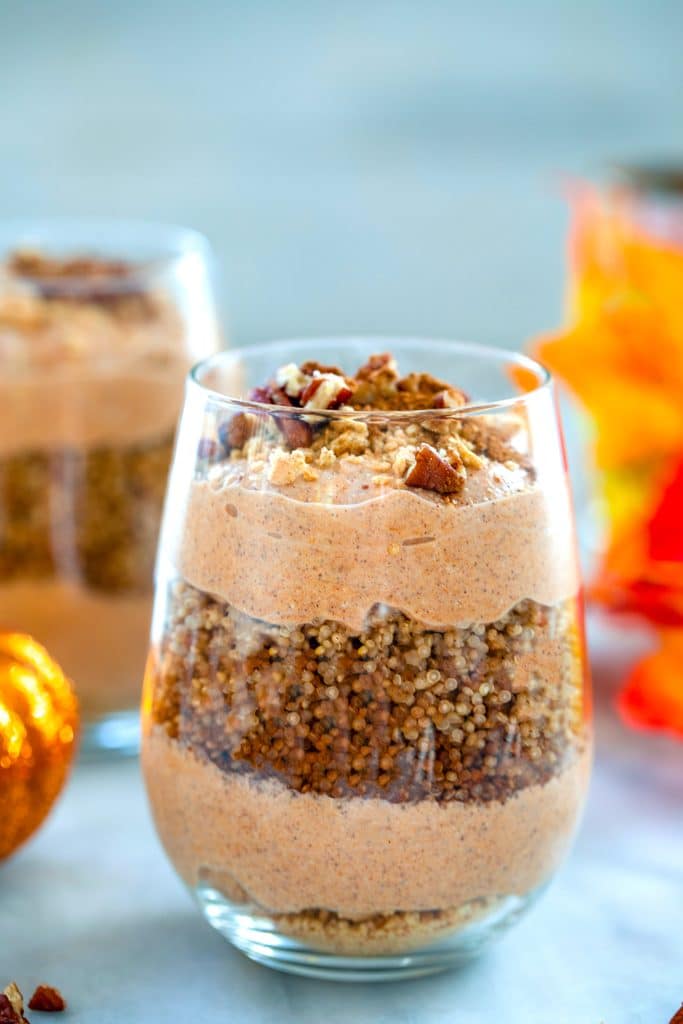 Head-on closeup view of pumpkin quinoa parfait with layers of graham cracker crumbs, pumpkin pie yogurt, and cinnamon quinoa with second parfait, glitter pumpkin and fall leaves in the background