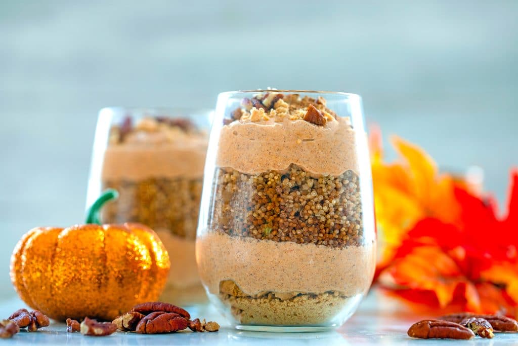 Landscape closeup head-on view of pumpkin quinoa parfait in a glass surrounded by crushed pecans, glitter pumpkin, fall leaves, and second parfait