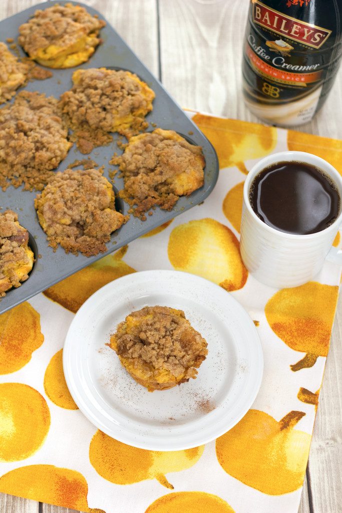 Bird's eye view of pumpkin spice french toast muffin on a white plate, muffin tin filled with muffins, cup of coffee, and bottle of coffee creamer on a pumpkin patterned towel