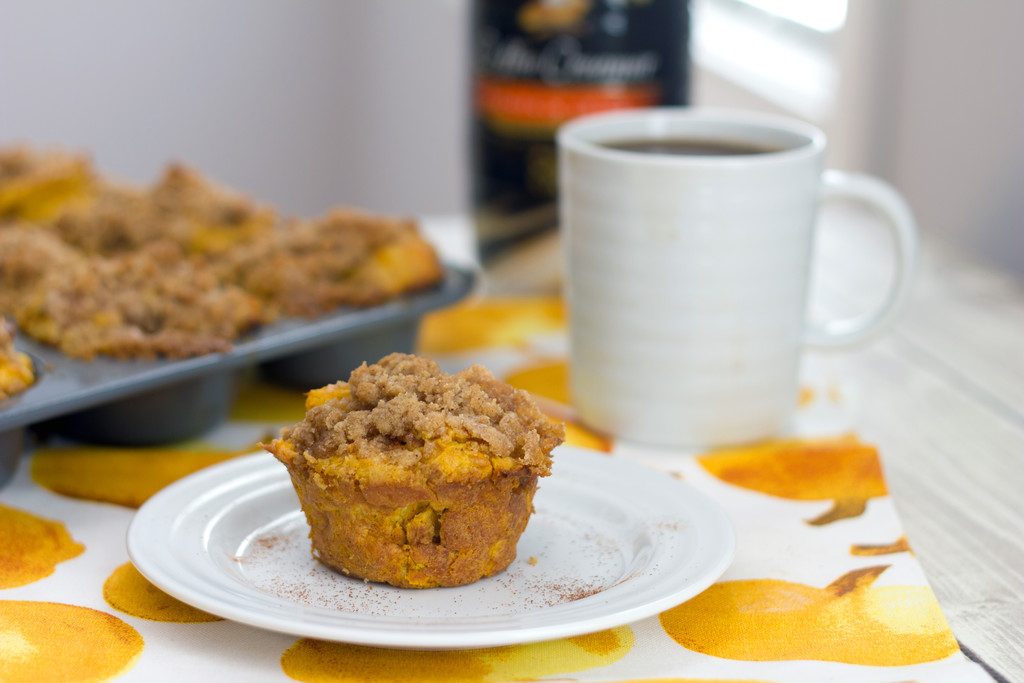 Landscape head-on view of a pumpkin spice french toast muffin on a white plate with muffin tin, cup of coffee, and bottle of coffee creamer in background