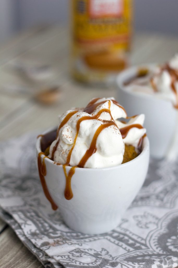 Head-on view of pumpkin spice latte mug cake topped with whipped cream with caramel drizzling down the sides and can of pumpkin and spices in the background