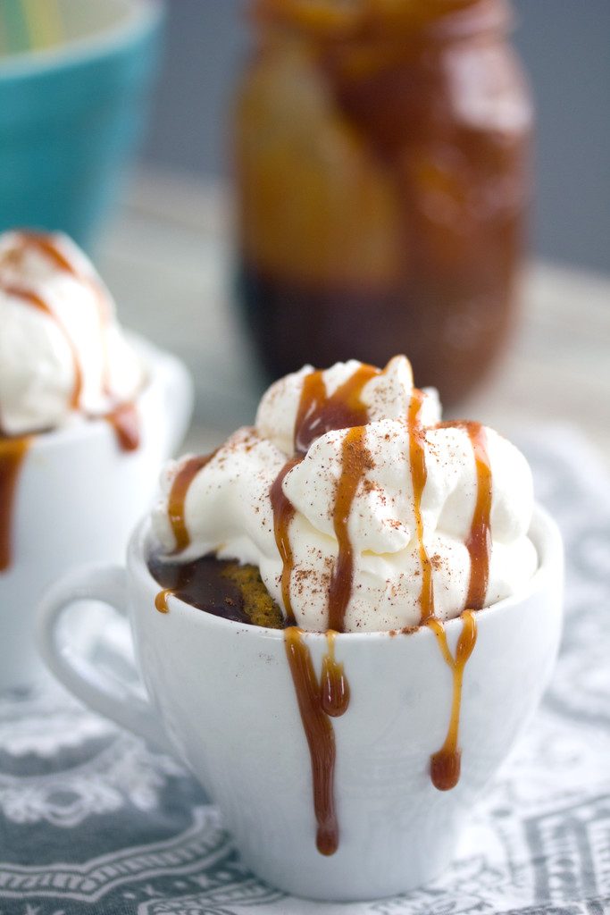 Head-on close-up view of pumpkin spice latte mug cake in a white mug with lots of whipped cream and caramel dripping down sides with second mug cake and jar of caramel in the background