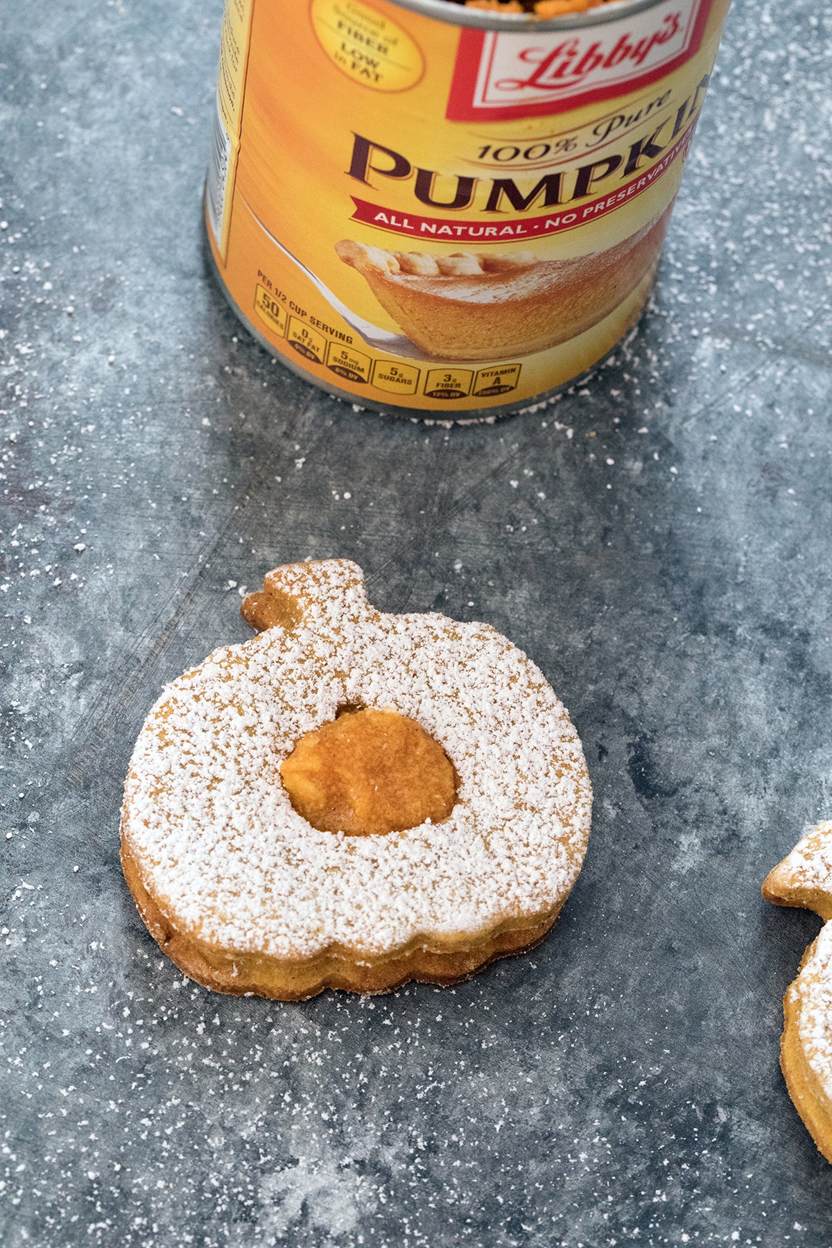 Overhead view of a pumpkin spice linzer cookie with confectioners' sugar and pumpkin filling on a dark surface with can of pumpkin puree in the background.