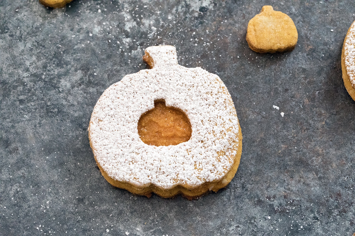 Landscape overhead closeup view of a pumpkin spice linzer cookie with confectioners' sugar and pumpkin filling on a dark surface.