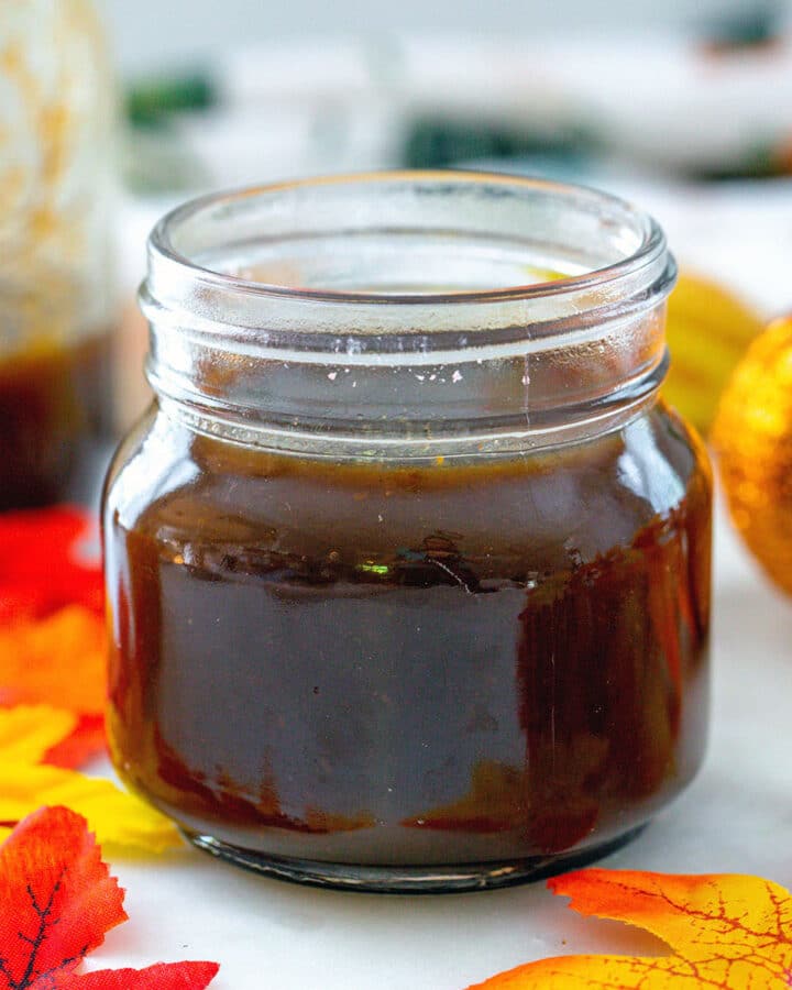 Head-on closeup view of a small jar of pumpkin spice syrup with fall leaves and large jar of syrup in background