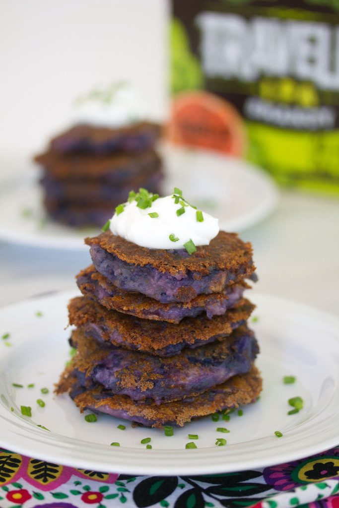 Head-on view of purple mashed potato pancakes in a stack and topped with Greek yogurt and chives with second plate of potato pancakes and box of beer in the background