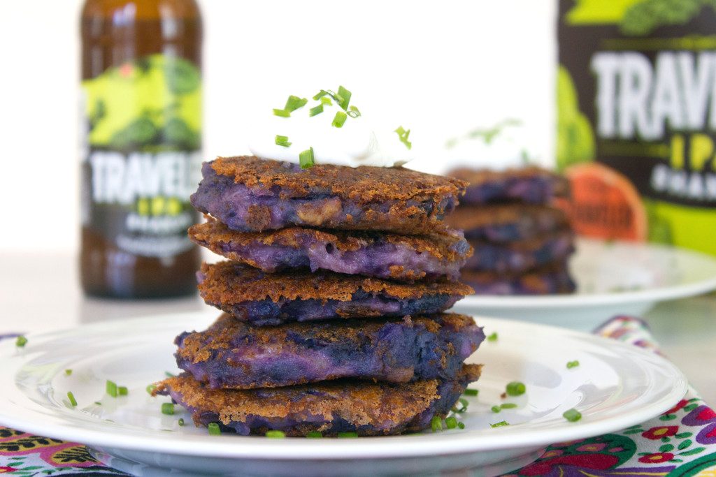 Landscape closeup view of a stack of purple mashed potato pancakes topped with Greek yogurt and chives with second plate of potato pancakes, bottle of beer, and box of beer in the background