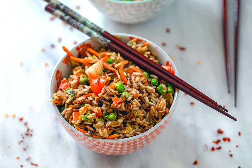 Landscape view of a bowl of quick vegetable fried brown rice with chopsticks balanced on top and red hot pepper flakes scattered around