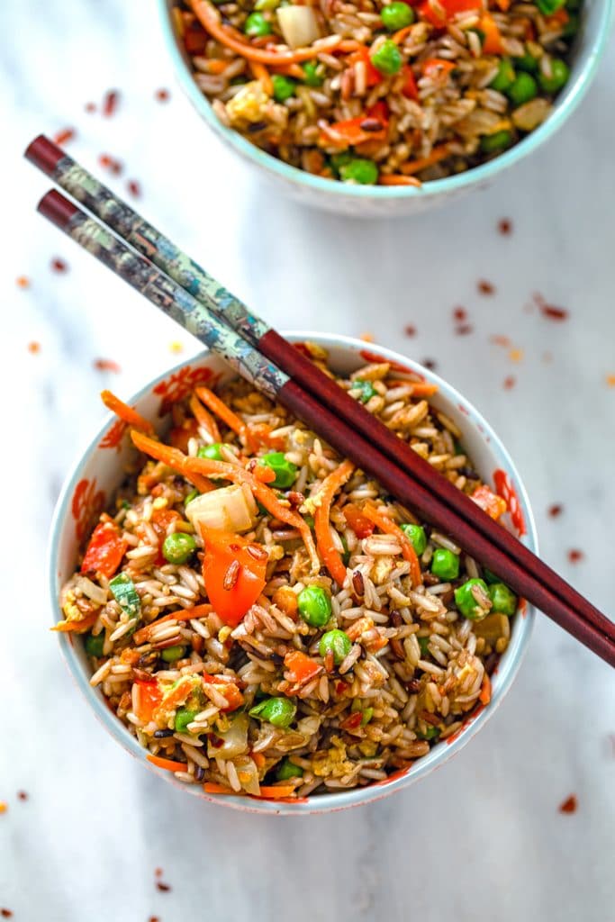 Overhead view of bowl of quick vegetable fried brown rice with chopsticks balancing on top and a second bowl in the background with red hot pepper flakes scattered around