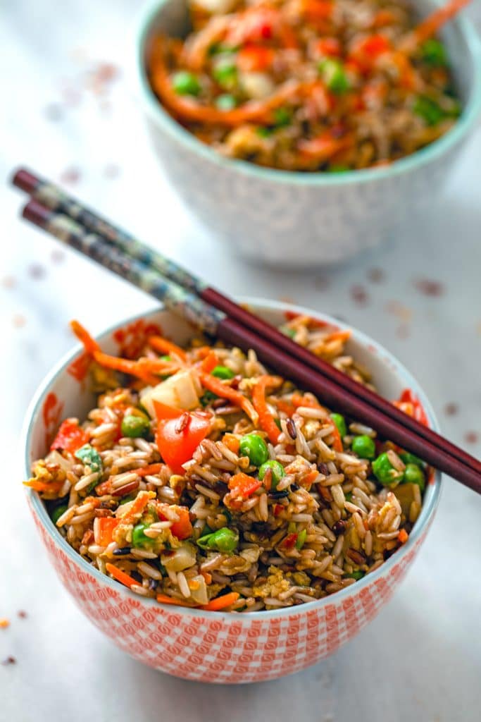 Head-on view of a bowl of quick vegetable fried brown rice with chopsticks balancing on it, a second bowl of rice in the background