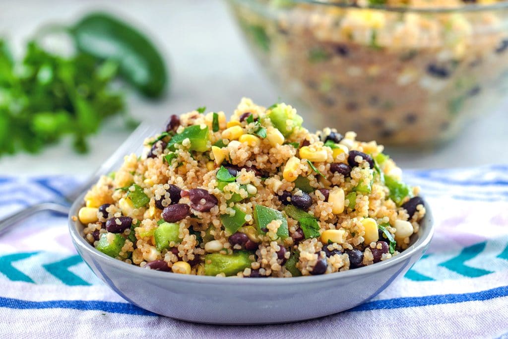 Landscape photo showing quinoa black bean and corn salad up close on purple towel with jalapeño, parsley, and large bowl of salad in the background