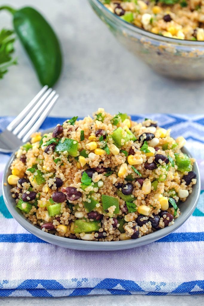 From above view of quinoa black bean corn salad on a colorful towel with fork, jalapeño, and large bowl of salad in the background