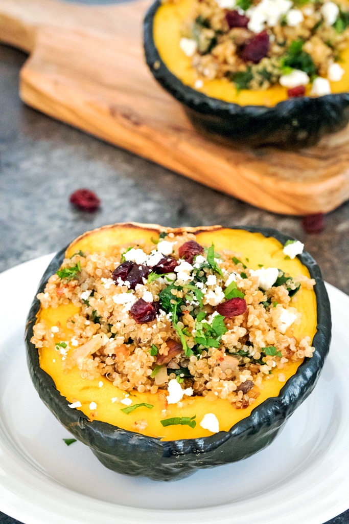 Overhead view of half an acorn squash stuffed with quinoa, spinach, feta, and dried cranberries on a white plate with second squash half on a cutting board behind it
