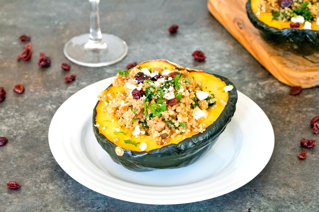 Landscape view of a white plate with half an acorn squash with quinoa, spinach, cranberries, and feta with wine glass and cutting board with second stuffed squash in the background and dried cranberries all around