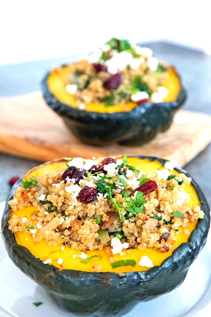 Head-on view of an acorn squash half overflowing with quinoa, spinach, feta, and dried cranberries with a second squash half on a cutting board in the background