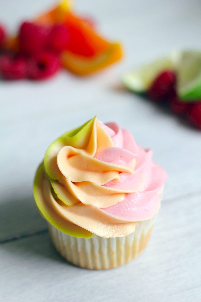 Overhead view of a rainbow sherbet cupcake with green, orange, and pink swirled frosting with raspberries, orange wedges, and lime wedges in background