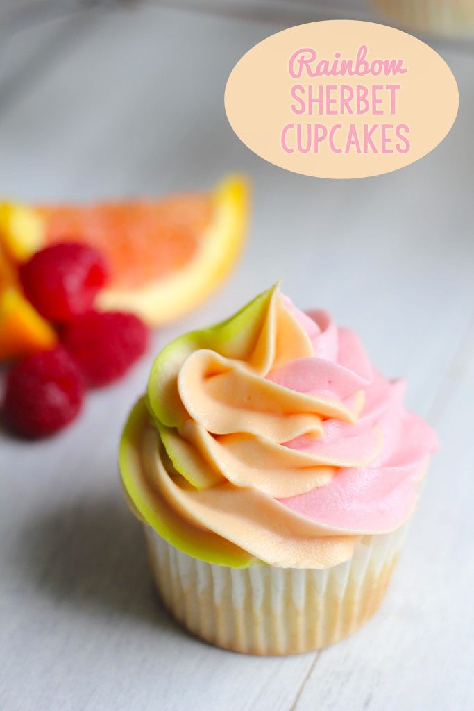 Head-on view of a rainbow sherbet cupcake with green, orange, and pink swirled frosting with raspberries and orange wedges in background and recipe title at top