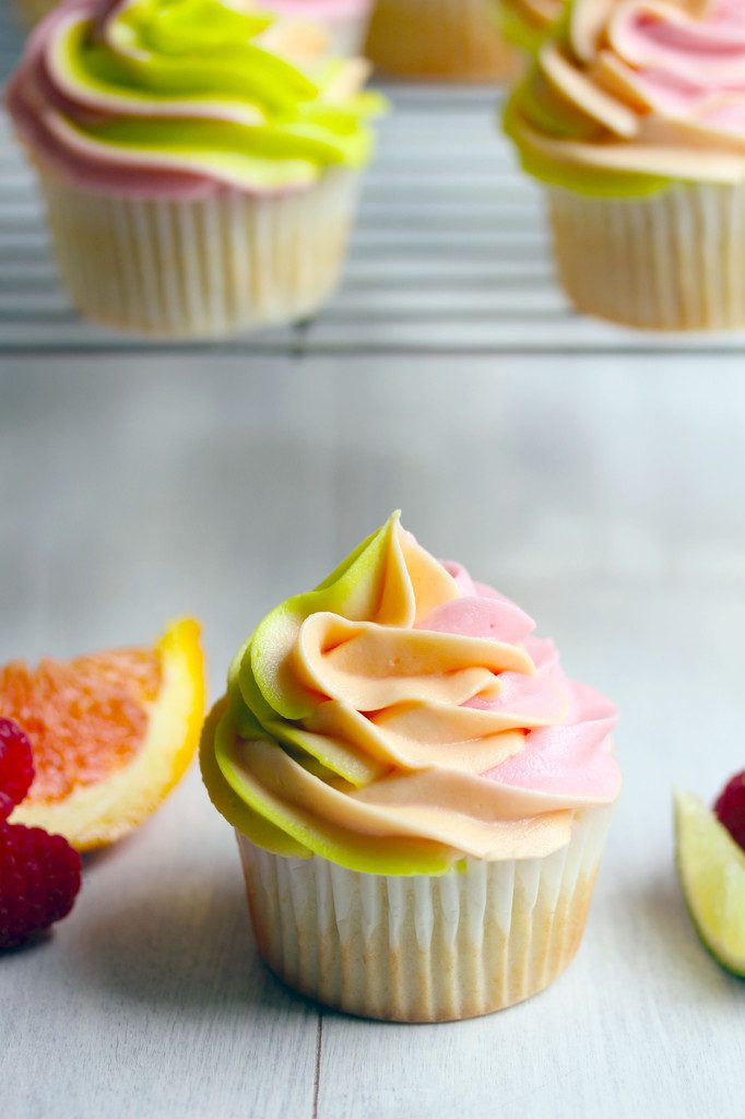 Head-on view of a rainbow sherbet cupcake with green, orange, and pink frosting with baking rack with more cupcakes in the background and orange wedges, lime wedges, and raspberries all around