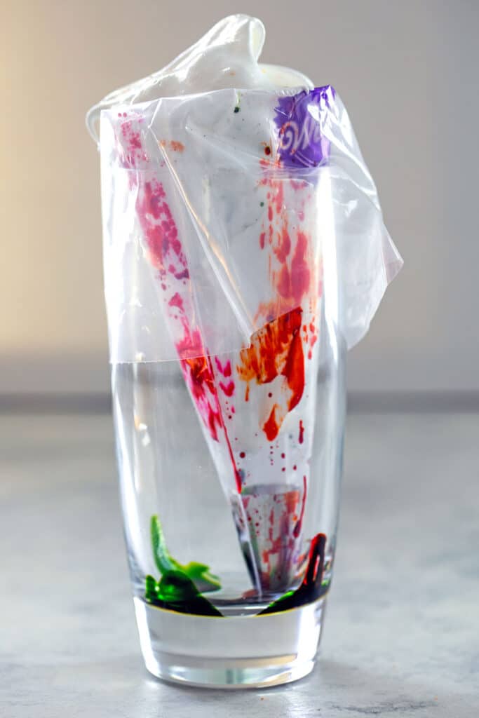 Head-on view of a tall drinking glass with a pastry bag in it and food coloring painted along the sides.