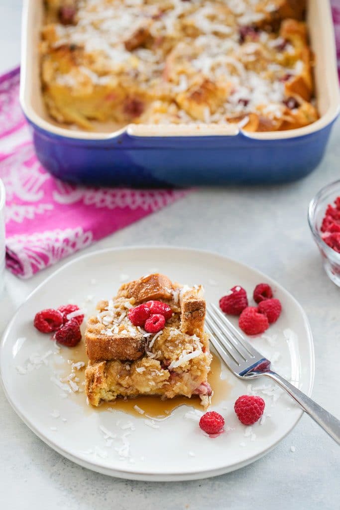 A serving of raspberry coconut french toast casserole, topped with fresh raspberries with a fork on the plate and with the baking dish on a pink dish towel in the background