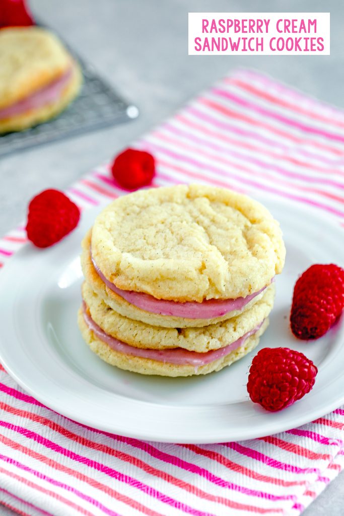 Head-on view of two raspberry cream sandwich cookies stacked on each other on a white plate on a pink striped towel with raspberries scattered around and recipe title at top