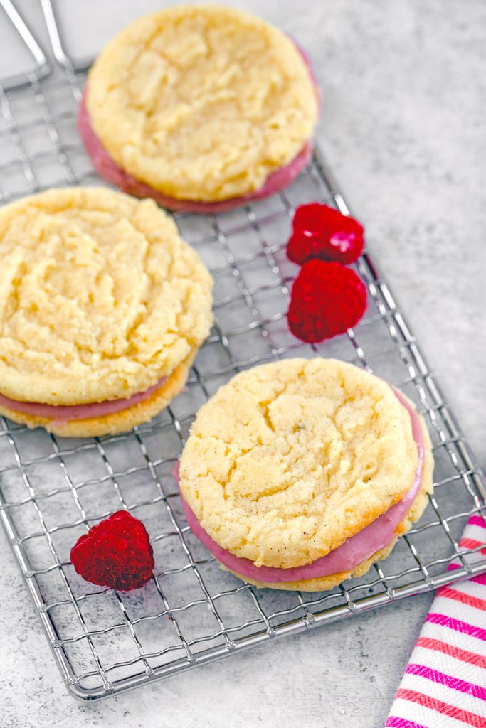 Overhead view of three raspberry cream sandwich cookies on a cooling rack with a few raspberries scattered around