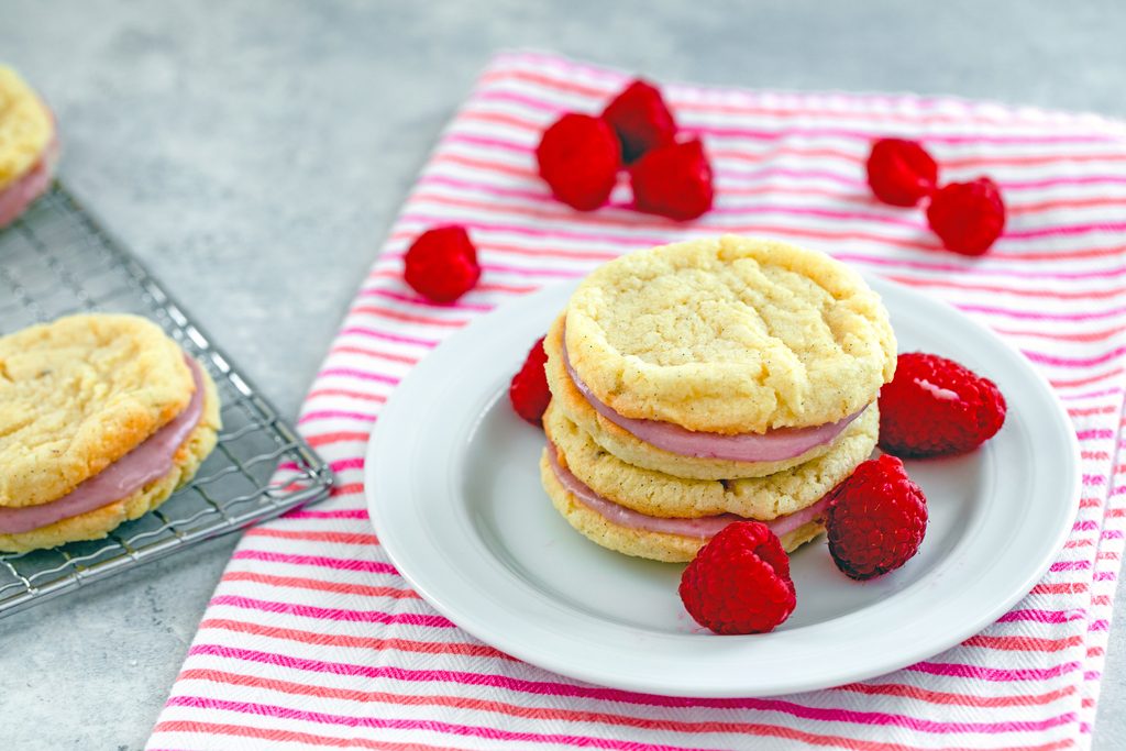 Landscape head-on view of two raspberry cream sandwich cookies on a white plate on a pink striped towel with cooling rack with more cookies and raspberries scattered around