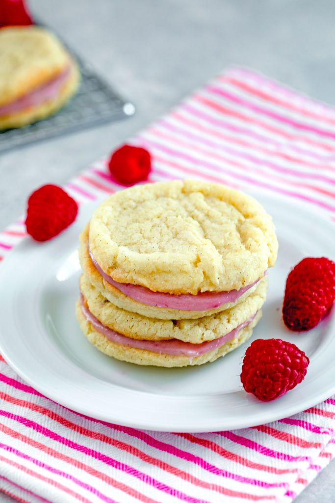 Head-on view of two raspberry cream sandwich cookies stacked on each other on a white plate on a pink striped towel with raspberries scattered around