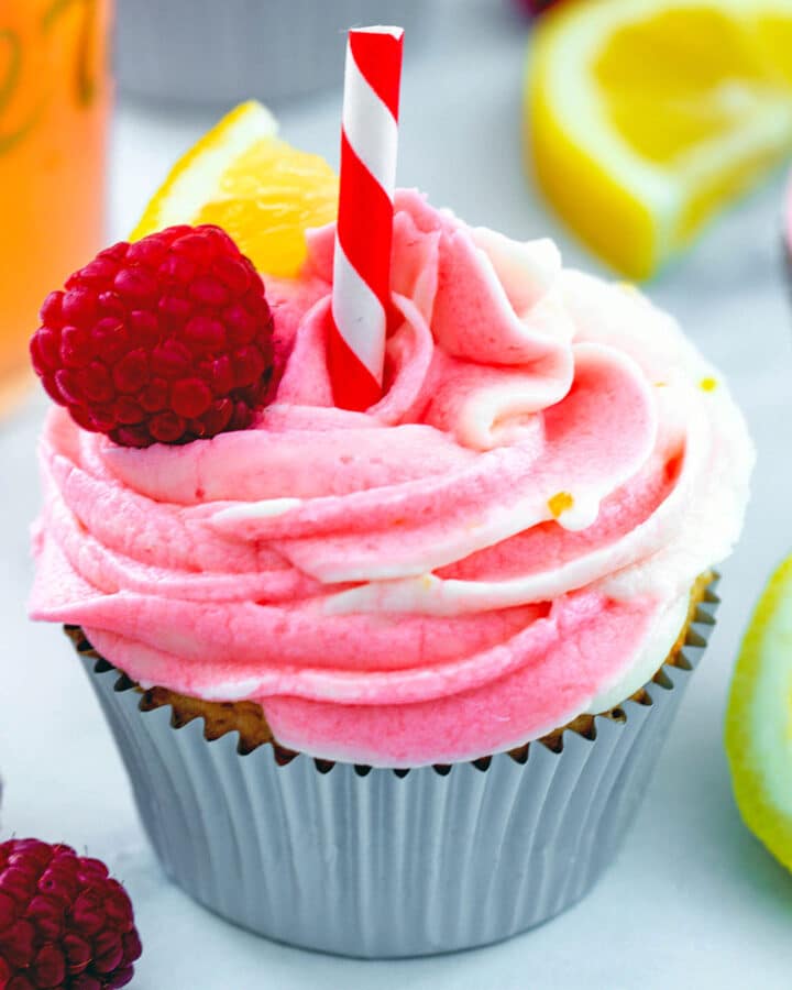 View of a raspberry lemonade cupcake with raspberry, lemon wedge, and straw garnish with raspberries and lemons in background